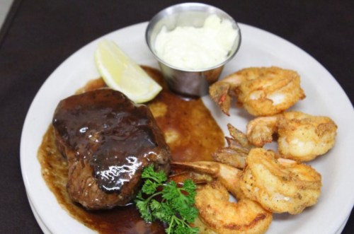A plate featuring a beef steak with sauce, fried shrimp, a lemon wedge, and a small cup of creamy sauce.
