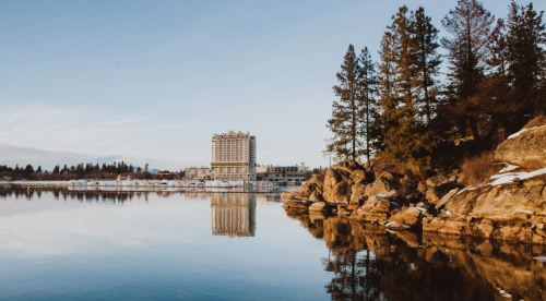 A serene lake scene with a hotel reflecting on the water, surrounded by trees and rocky shoreline under a clear sky.