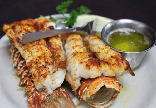 Grilled lobster tails seasoned and served with melted butter and a slice of lemon on a white plate.