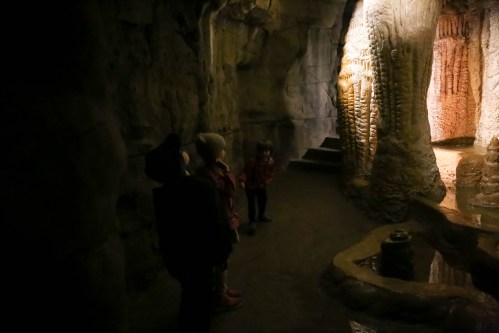 Three children explore a dimly lit cave, with rock formations and a small water feature in the background.