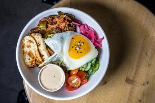 A colorful bowl featuring a fried egg, vegetables, pickled onions, and a creamy sauce on a wooden table.