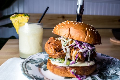 A close-up of a sesame seed burger with fried chicken, slaw, and sauce, alongside a tropical drink with a straw.
