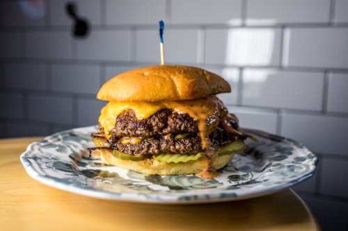 A juicy double cheeseburger with melted cheese, pickles, and a toothpick, served on a decorative plate.