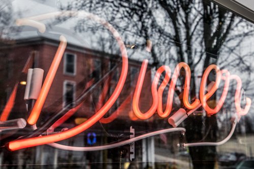 Neon sign reading "Diner" in bright red, reflecting in a window with trees and a building in the background.