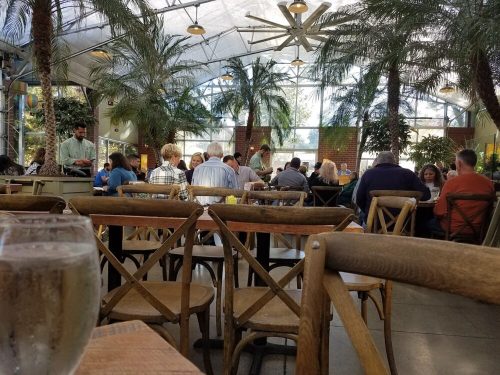 A bustling indoor restaurant with palm trees, filled with diners at wooden tables and chairs.