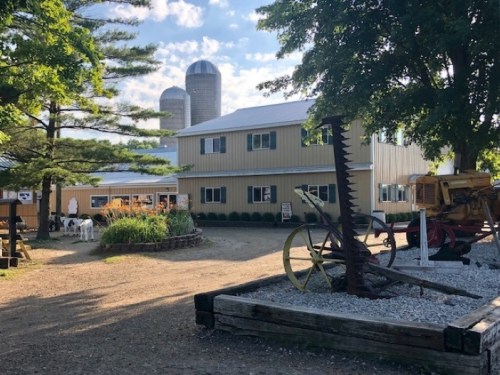 A farm scene featuring a barn, silos, and vintage farm equipment surrounded by trees and gravel pathways.