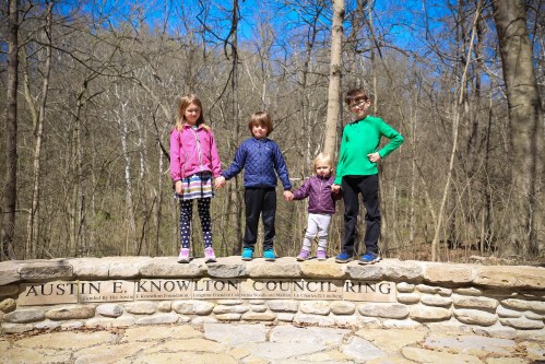 Four children stand on a stone wall in a wooded area, holding hands and smiling, with trees and blue sky in the background.