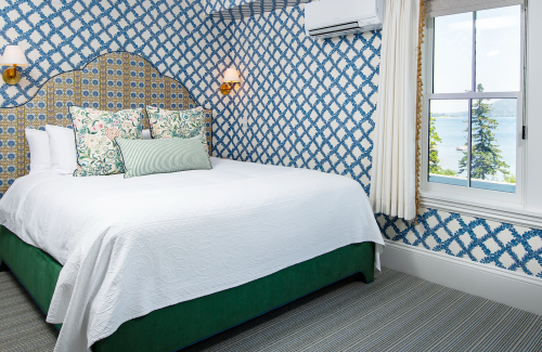 Cozy bedroom with a patterned wall, a white bedspread, decorative pillows, and a window overlooking a scenic view.