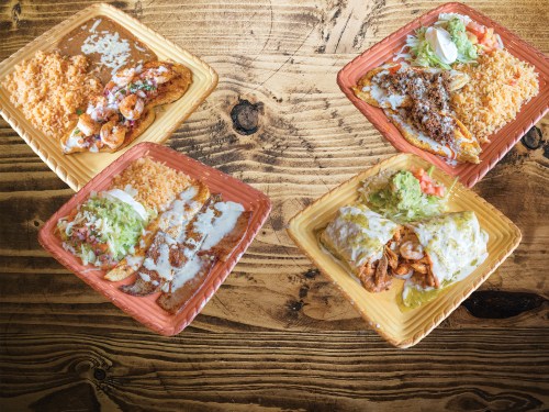 Four colorful plates of Mexican food featuring shrimp, rice, tacos, and toppings on a wooden table.