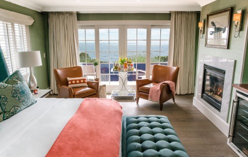 Cozy hotel room with a bed, two armchairs, a fireplace, and large windows showcasing a scenic ocean view.
