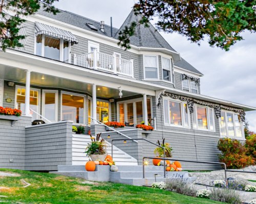 A charming gray house with a wraparound porch, decorated with pumpkins and flowers, set against a cloudy sky.