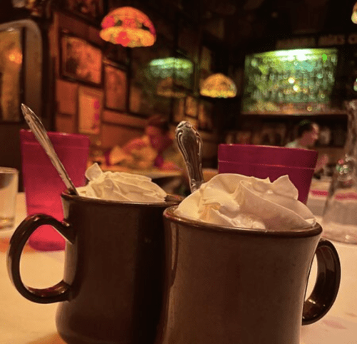 Two mugs with whipped cream sit on a table, surrounded by colorful lighting and a cozy restaurant atmosphere.