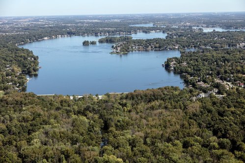 Aerial view of a serene lake surrounded by lush greenery and residential areas, under a clear blue sky.