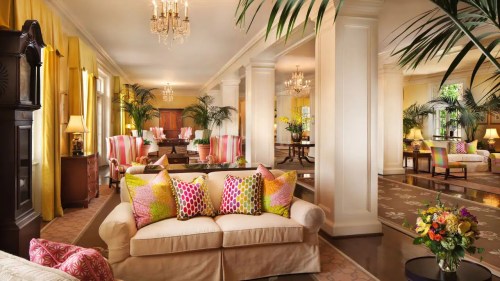 Bright and elegant living room with plush sofas, colorful pillows, and lush plants, featuring chandeliers and stylish decor.
