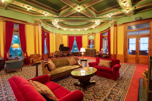 Elegant lounge with red curtains, plush seating, and a grand piano, featuring ornate ceiling and decorative furnishings.