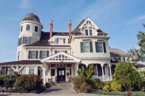 A large, elegant Victorian-style house with a turret, surrounded by lush greenery and colorful flowers.
