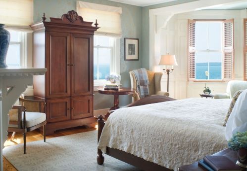 Cozy bedroom with a wooden armoire, armchair, and ocean view through large windows. Soft lighting and elegant decor.