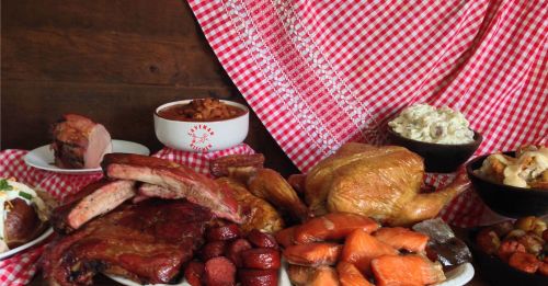 A spread of various meats, including ribs, chicken, and sausage, alongside sides like potato salad and baked beans on a checkered tablecloth.