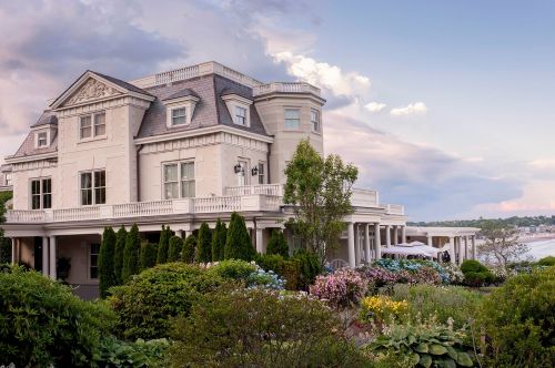 A large, elegant mansion surrounded by lush gardens and a scenic view of the water under a cloudy sky.
