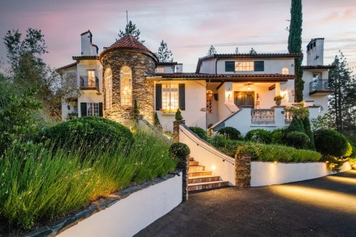 A charming stone and stucco house with a circular tower, surrounded by lush greenery and softly lit at dusk.