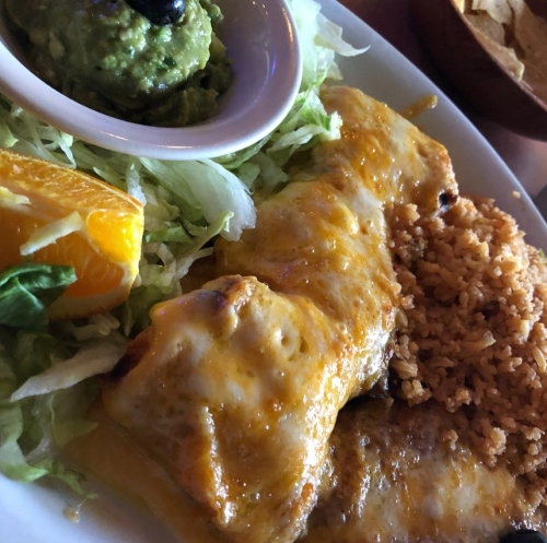 A plate of enchiladas topped with melted cheese, served with rice, guacamole, lettuce, and an orange slice.