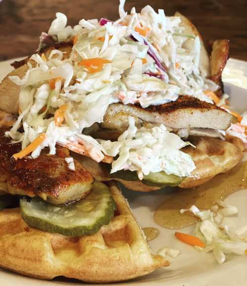 A plate of waffles topped with grilled chicken, coleslaw, and pickles, drizzled with syrup.