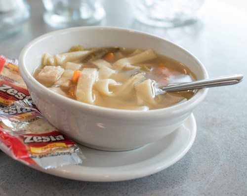 A bowl of chicken noodle soup with vegetables, served with two packets of Zesta crackers on the side.