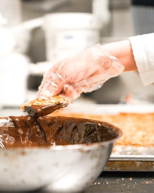 A gloved hand dips a piece of dessert into a bowl of melted chocolate in a kitchen setting.