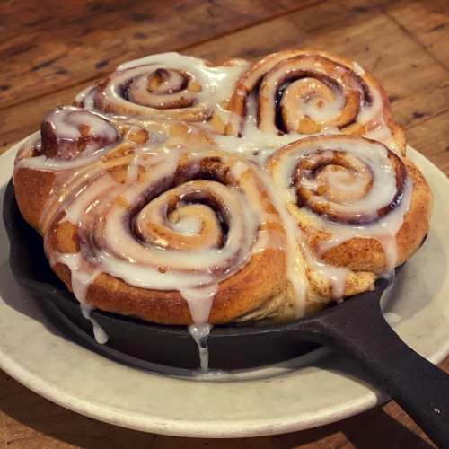 A freshly baked cinnamon roll in a cast iron skillet, drizzled with creamy icing, on a wooden table.