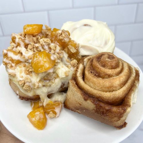A plate with two cinnamon rolls: one topped with cream cheese and the other with peaches and granola.