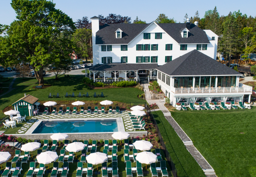 Aerial view of a white hotel with a pool and lounge chairs, surrounded by green grass and trees on a sunny day.