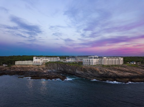 A modern hotel sits on rocky cliffs by the ocean, under a colorful sunset sky with clouds.
