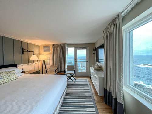 Cozy hotel room with a bed, ocean view, and modern decor, featuring a balcony and large windows.