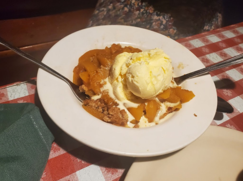 A bowl of peach cobbler topped with a scoop of vanilla ice cream, served with two spoons on a checkered tablecloth.