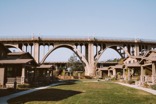 A large concrete bridge arches over a grassy area with small houses beneath it, under a clear blue sky.