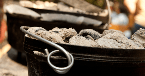 Close-up of a cast iron Dutch oven covered with ash and coals, ready for outdoor cooking.