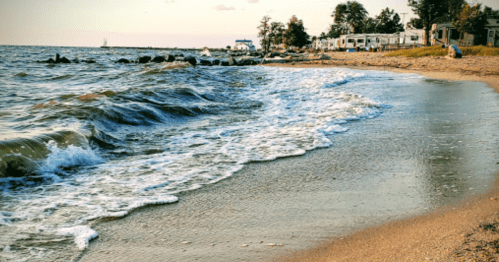Waves gently lap at a sandy beach, with rocky outcrops and trees in the background under a clear sky.