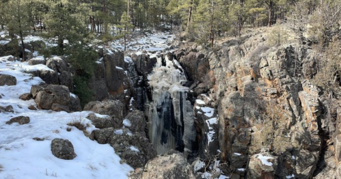 A waterfall cascades over rocky terrain, surrounded by pine trees and patches of snow.