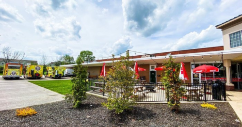 A food truck area with picnic tables, red umbrellas, and greenery under a cloudy sky.