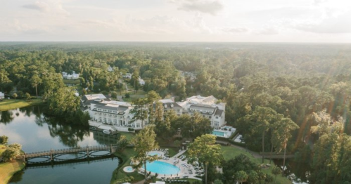 Aerial view of a luxurious hotel by a lake, surrounded by lush greenery and a swimming pool.