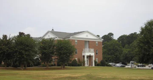 A brick building with white accents, surrounded by trees and a grassy area, under a cloudy sky.