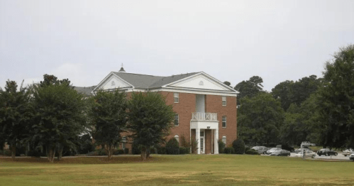 A brick building with white accents, surrounded by trees and a grassy area, under a cloudy sky.