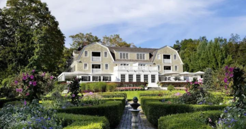 A large, elegant house surrounded by lush gardens and colorful flowers under a clear blue sky.