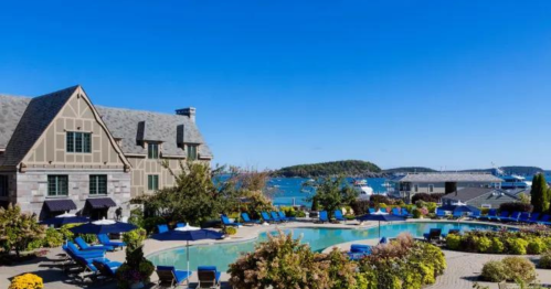 A scenic view of a hotel with a pool, surrounded by lounge chairs and lush greenery, overlooking a harbor and blue sky.