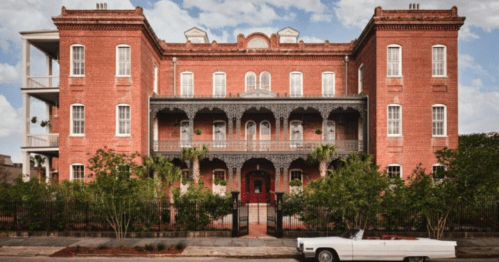 Historic red brick building with ornate balconies and lush greenery, featuring a classic white convertible parked in front.