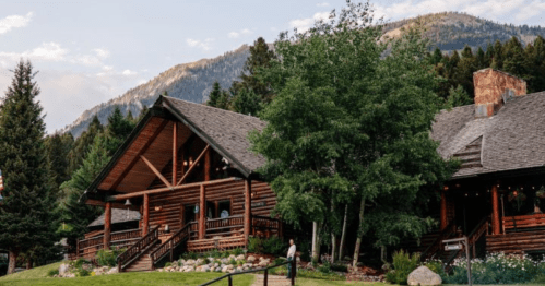 A rustic log cabin surrounded by trees and mountains, with a clear sky and outdoor seating area.