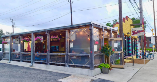 A cozy restaurant with outdoor seating, string lights, and colorful decor, surrounded by power lines and greenery.