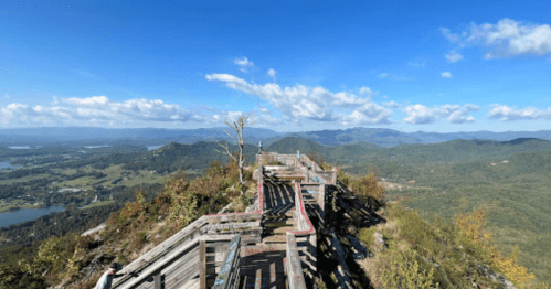 A panoramic view from a mountain overlook, featuring wooden railings and lush green valleys under a blue sky.