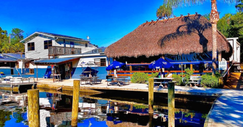 A waterfront restaurant with a thatched roof, surrounded by boats and palm trees, reflecting in calm water.