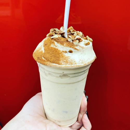 A hand holding a creamy milkshake topped with crushed nuts and a sprinkle of cinnamon against a red background.